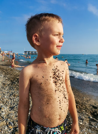 Beautiful boy squints his eyes from the sun, looks up into the sky on the beach by the sea. Stuck seas sand on the bodyの写真素材