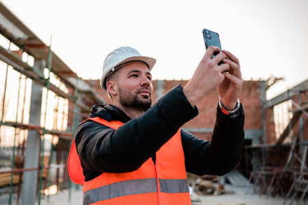 Civil engineer or architect with hardhat and orange construction vest on construction site taking photos of the progress with the smartphone camera for the report.の写真素材