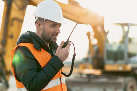 Young engineer talking radio communication (walkie talkie) and wearing a white helmet and construction orange vest. Close up engineers working on a building site with the sunny background.の写真素材