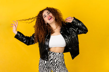 Funny and cool girl with curly hair in black leather jacket posing. Isolated on yellow background.