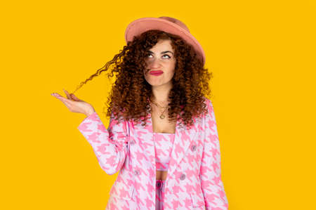 Girl with curly hair in summer pink outfit with hat smiling and enjoying. Posing on the yellow background.の写真素材