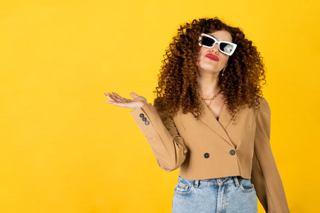 Curly girl in the trendy outfit of jeans and jacket posing on the yellow background in the studio. Woman facial expression. Isolated portrait.の写真素材
