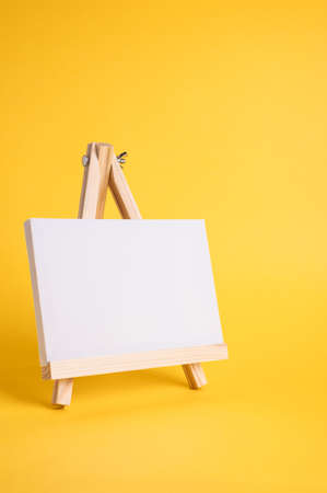 White canvas on a small wooden easel stand minimal concept on the yellow backdrop. Empty blank mock up frame for design with copy space.の写真素材