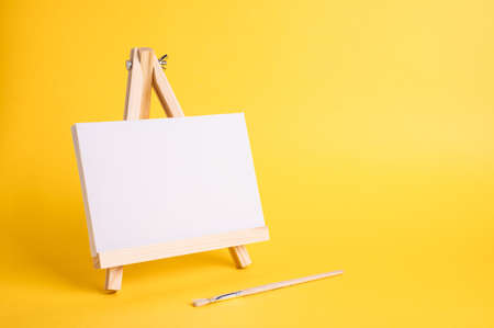 White canvas on a small wooden easel stand minimal concept on the yellow backdrop. Empty blank mock up frame for design with copy space.の写真素材
