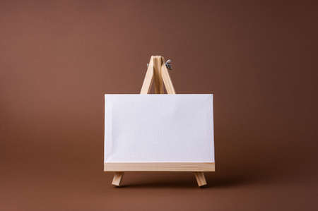White canvas on a small wooden easel stand minimal concept on the brown backdrop. Empty blank mock up frame for design with copy space.の写真素材