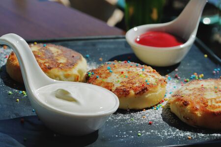 cheese pancakes with sour cream and lies on a square black plate in a dark smoky background with warm lighting. Beautiful greens on the background. Close up. Spaceの写真素材
