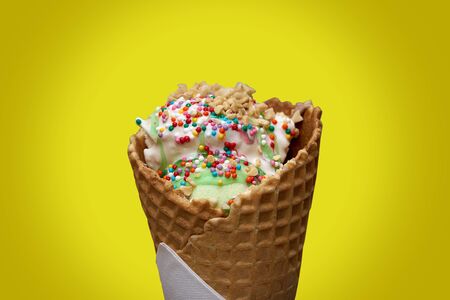 white ice cream in a waffle cone with colored sprinkles and pieces of nuts yellow backgroundの写真素材