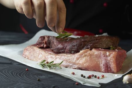 Chef salts steak with herbs on paper. Preparing fresh beef or pork. Horizontal photo with a dark black background.の写真素材