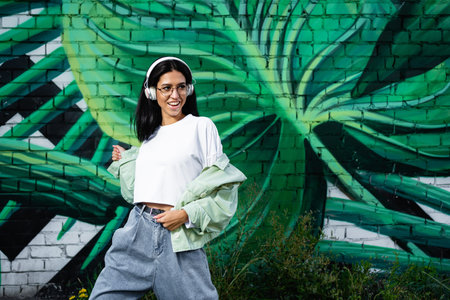 A brunette girl in glasses and white headphones is dancing against the background of a wall with...