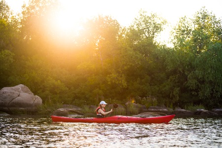 Young Woman Paddling the Red Kayak in Beautiful Lagoon with Green Trees and Stones at Warm Summer Sunsetの写真素材