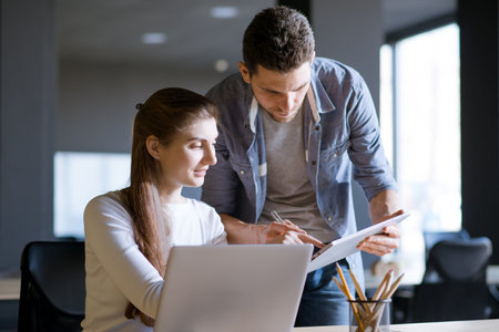 Young Smiling Coworkers Woman and Man Discusing Working Details and Pointing at Laptop Screen in the...