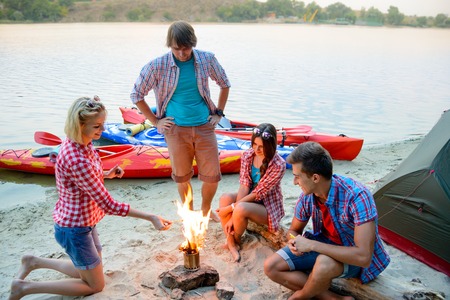 Young Happy Travelers Resting at the Evening near Fire on the Sand Beach Infront of Kayaks, Tent and River. Travel, Adventure and Water Recreation Concept.の写真素材