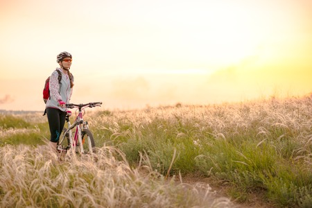 Young Woman Riding the Mountain Bikes in the Beautiful Field Full of Feather Grass at Sunset. Adventure and Travel Concept.の写真素材