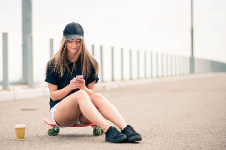 Young Beautiful Smiling Blonde Girl Using Smartphone while Sitting on the Skateboardの写真素材