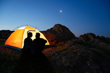 Young Couple Sitting near Illuminated Tent and Looking at Each Other at Beautirul Evening in the Mountainsの写真素材