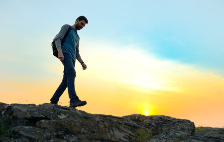 Young Happy Man Traveler Hiking with Backpack on the Rocky Trail at Warm Summer Sunset. Travel and...