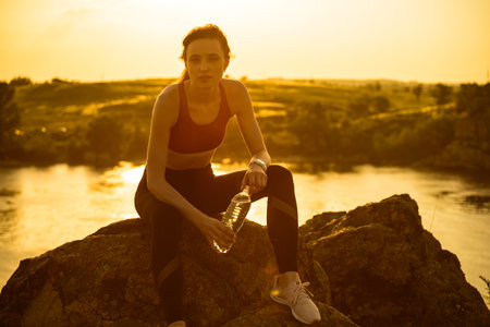Woman Resting and Drinking Water after Running Outdoor. Workout at Hot Summer Sunset. Sport and...