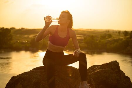 Woman Resting and Drinking Water after Running Outdoor. Workout at Hot Summer Sunset. Sport and Healthy Active Lifesyle.の写真素材