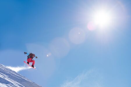 Snowboarder Jumping on the Red Snowboard in the Mountains in Bright Rays of Sun. Freeride Snowboarding and Extreme Winter Sportsの写真素材