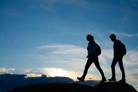 Couple of Young Happy Travelers Hiking with Backpacks on the Beautiful Rocky Trail at Warm Summer Sunset. Family Travel and Adventure Concept.の写真素材