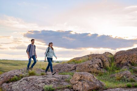 Couple of Young Happy Travelers Hiking with Backpacks on the Beautiful Rocky Trail at Warm Summer Sunset. Family Travel and Adventure Concept.の写真素材