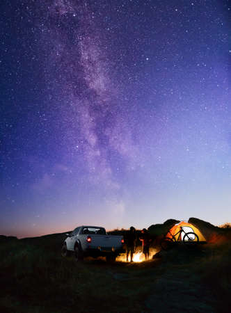 Friends near Bonfire, Pickup Truck, Tent and Bike in the Mountains under Night Sky with Milky Way. Adventure and Travelの写真素材