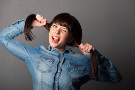 Portrait of young pretty cute girl holding her brown hairs and showing her tongue on grey の写真素材