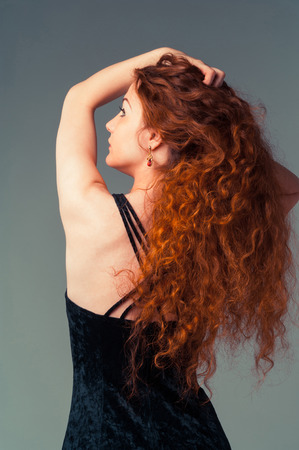 Portrait of young beautiful woman in black dress with long red hairs standing on grey backgroundの写真素材