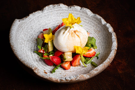 A vibrant salad showcases a creamy burrata cheese at its center, surrounded by fresh strawberries, greens, and edible flowers, elegantly presented on a textured, rustic plate.