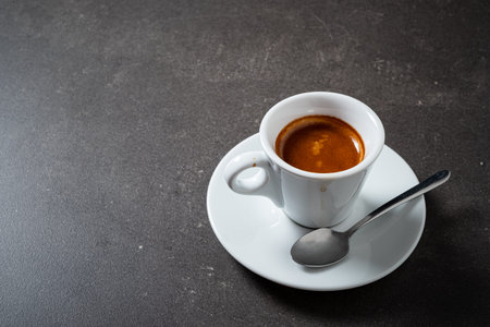 A small white cup filled with dark, aromatic espresso, resting on a matching saucer, with a silver...