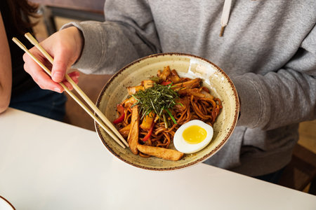 A person expertly holds a bowl of tasty noodles topped with veggies and a soft-boiled egg.の写真素材