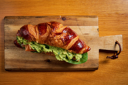 Croissant filled with creamy avocado, juicy tomato, and crisp greens on a wooden board.の写真素材