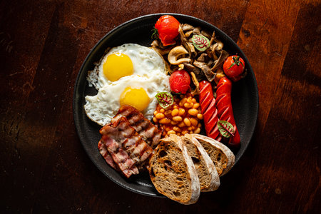 Tasty breakfast with sunny-side-up eggs, crispy bacon, sausages, and veggies on a dark plate.の写真素材