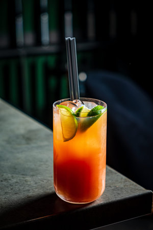 Brightly colored cocktail filled with lime wedges sits on a sleek bar countertop at night.