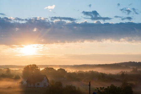 Golden rays of sunlight break through clouds, illuminating a quiet rural scene filled with mist.の写真素材
