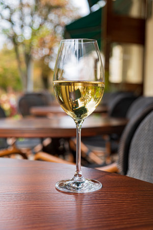 A glass of white wine sits elegantly on a wooden table, surrounded by trees and outdoor seating.の写真素材