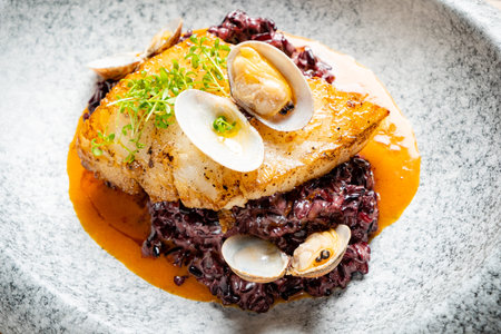 A beautifully plated seafood dish features tender fish, black rice, and fresh clams.