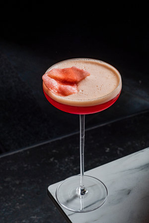A pink cocktail in a stemmed glass rests on a counter, garnished with chopped fruit.の写真素材