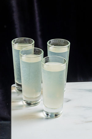 Four shot glasses are filled with pale lemonade. They stand on a marble surface.