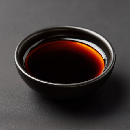 A small black bowl holds soy sauce, reflecting deep hues against a dark surface, inviting taste.の素材