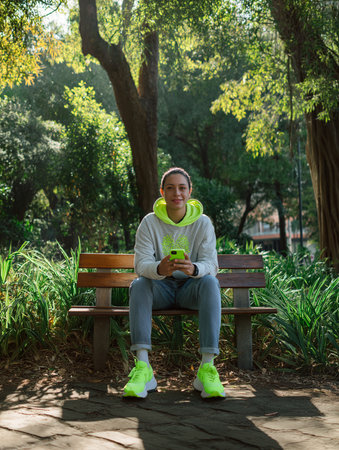 A Latino person sits on a bench, smiling and using a smartphone in a green park setting.の素材