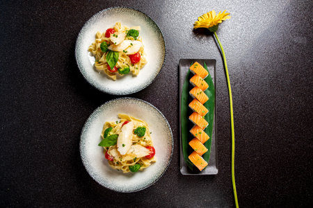 Two plates of creamy pasta and vegetables, plus sushi on a sleek tray.の写真素材