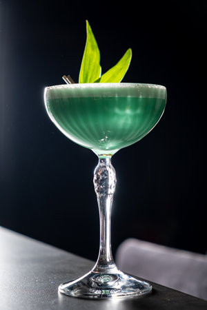 A green cocktail rests elegantly in a stylish glass on the bar, framed by a dark backdrop.の写真素材