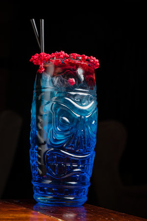 A colorful tiki drink sits on a wooden bar top, showcasing a blue hue topped with red garnishes.の写真素材