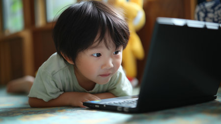 A joyful boy is playing on a laptop, lying on the floor in a bright and inviting room.の素材