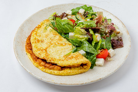 Fluffy omelet with melted cheese sits beside a vibrant salad of greens, tomatoes, and feta.の写真素材