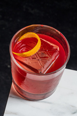 An elegant cocktail in a clear glass, with a large ice cube and an orange twist.