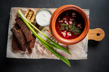 A colorful bowl of borscht with bread, garlic, and green onions on a rustic board.