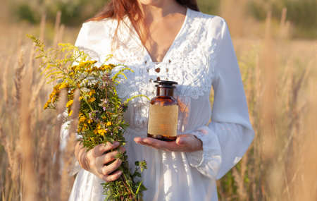 A woman in a white dress is holding a brown glass bottle and a bouquet of herbs. A blank label with space for a description. Alternative Medicine and natural remedies. The idyllic atmosphere of the photo.の写真素材
