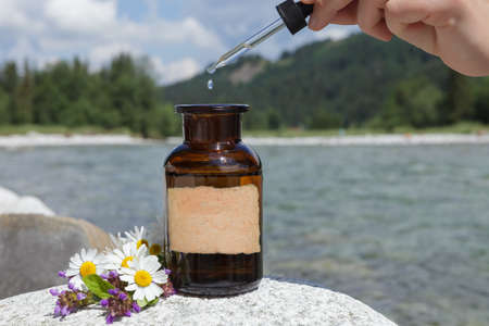 Conceptual image related to natural medicine. Drip from dropper falling into bottle. Old medical bottle against background of mountains. Empty label, mockup /の写真素材